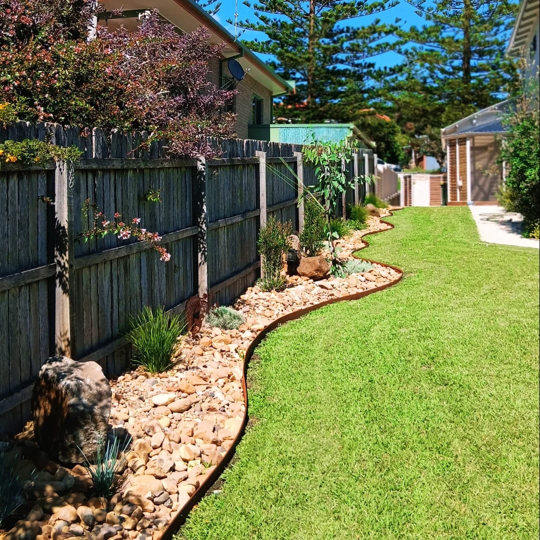 A sweeping river pebble bed with steel edging curving alongside a lawn, planted with grasses and shrubs against a timber fence