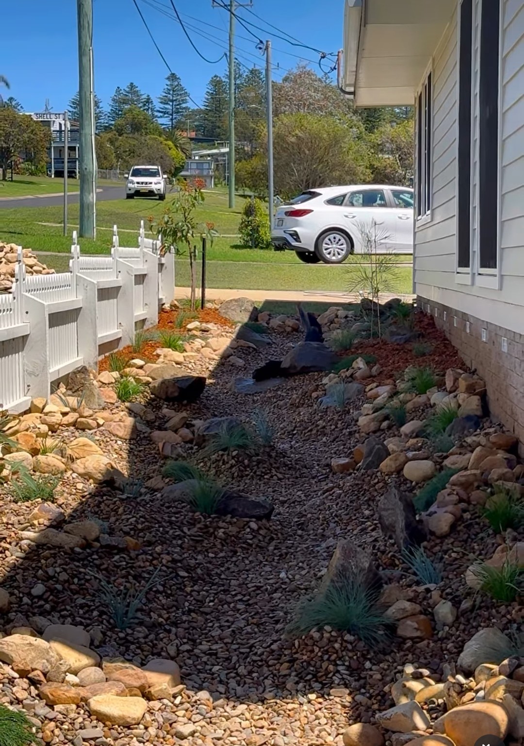 A finished dry creek bed of river stones and feature rocks running between a house and a white picket fence, planted with tufting grasses